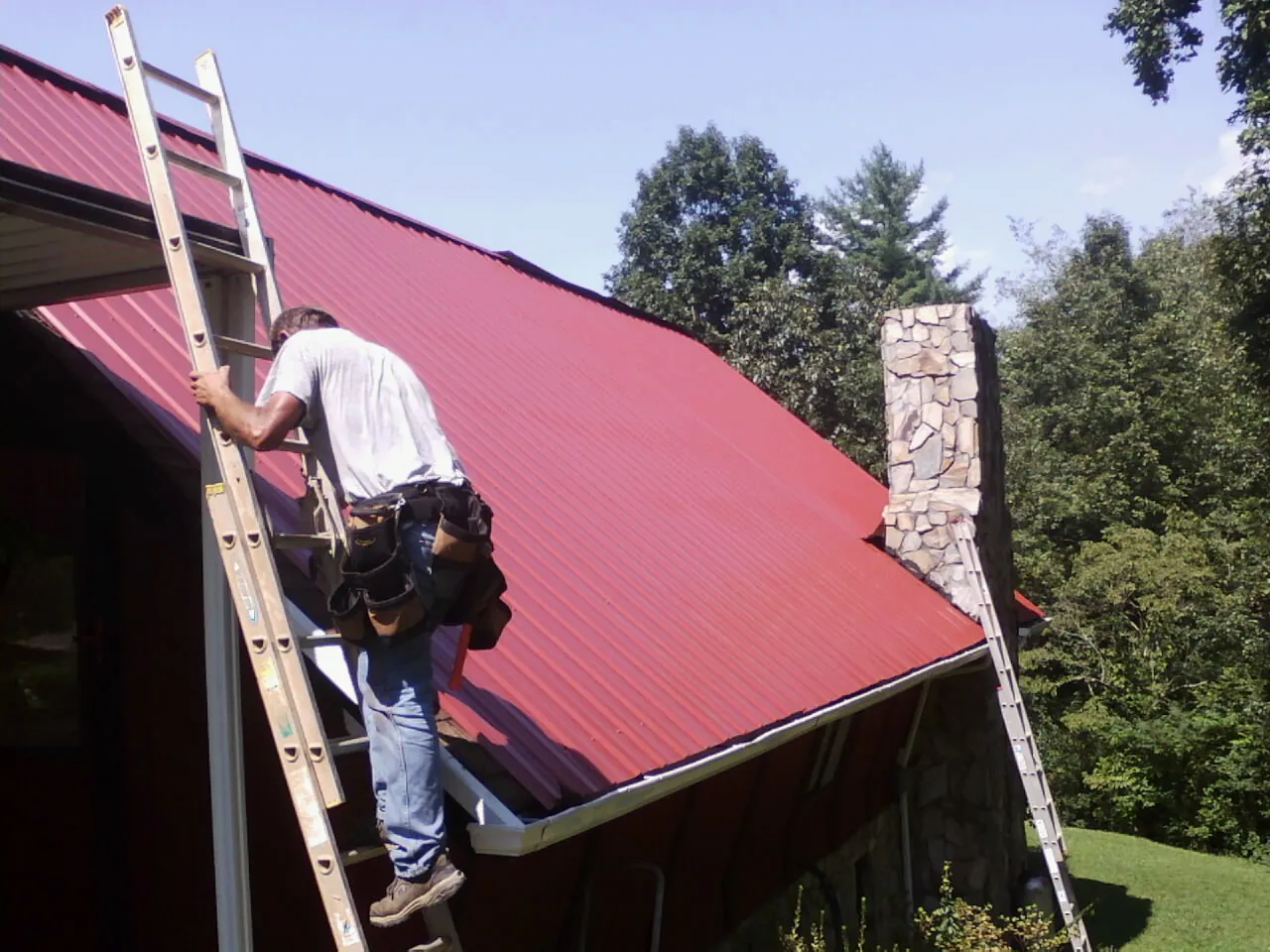 Roofer on a ladder beside a red metal roof and stone chimney — Soffit & Fascia Repair by Roofing Brothers in Greenport